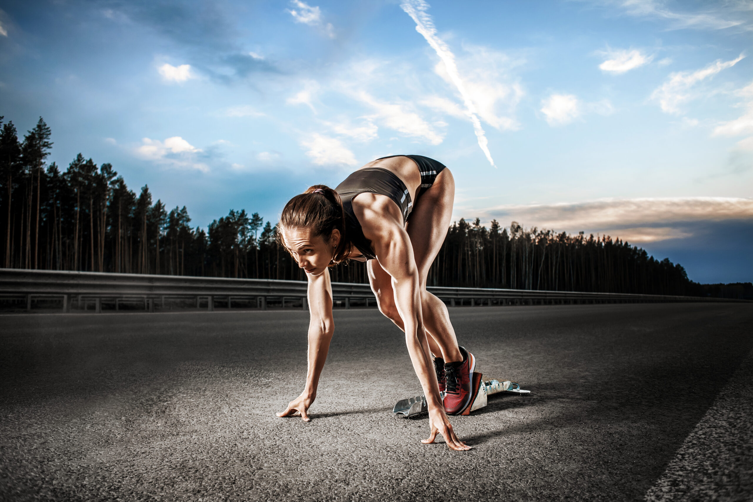 Athlete in a sprinting position on an empty road, preparing to start a race. Lush trees and a vibrant sky in the background enhance the dynamic scene.
